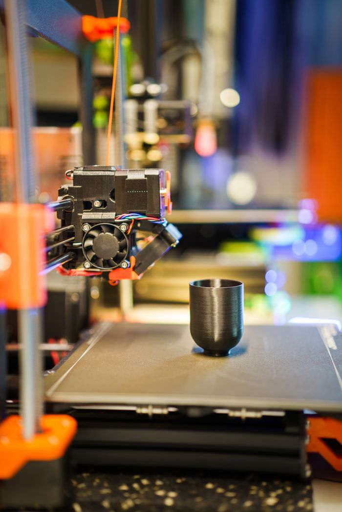 services-02 Detailed shot of a 3D printer manufacturing a black plastic cup indoors.
