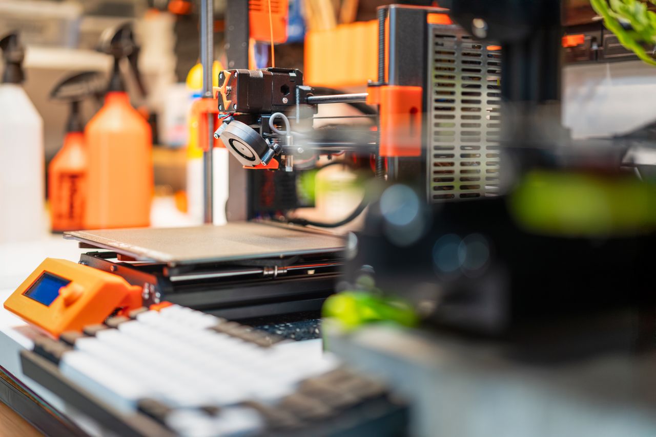 gallery-4 Detailed view of a 3D printer in a busy workshop, showcasing modern 3D printing technology and equipment.